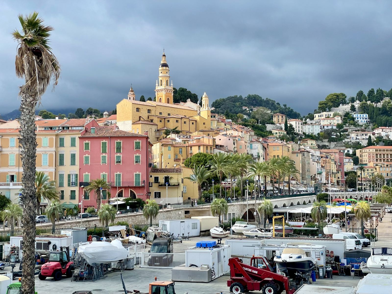 Menton from the harbour — pastel-coloured buildings, church tower, palm trees, stormy sky